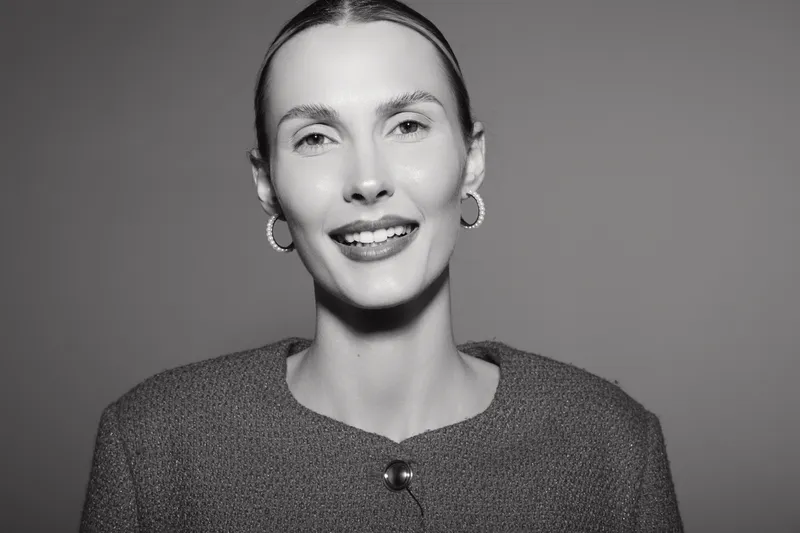 Smiling Caucasian woman with fair skin and slicked back hair wears hoop earrings and textured jacket. Studio portrait on a plain background with soft lighting and positive mood.