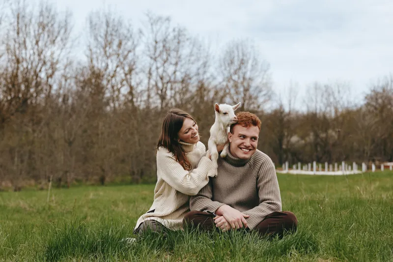 A couple enjoying a relaxed moment outdoors in a grassy field with a playful goat kid nearby. Warmth and companionship define this lifestyle scene, capturing nature, joy, and gentle animal