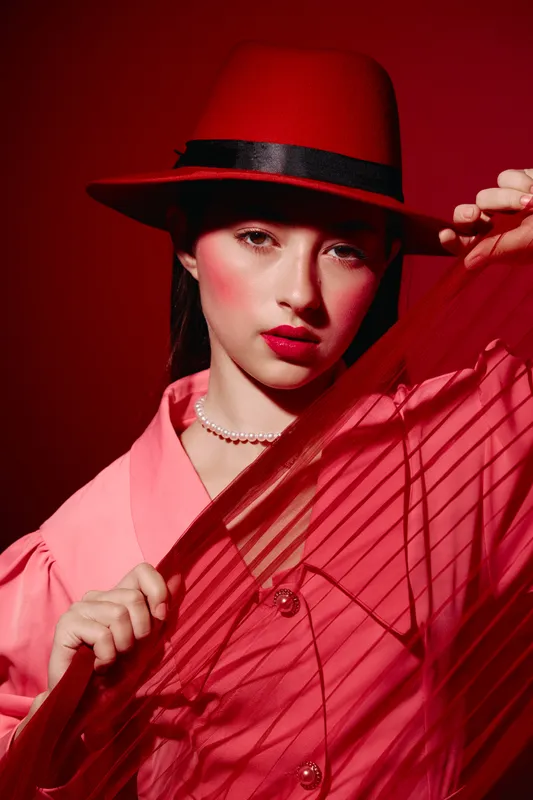Fashionable young woman in a red hat and pink blouse poses with enthusiasm. Dramatic shadows create a bold and creative beauty concept with warm lighting mood.