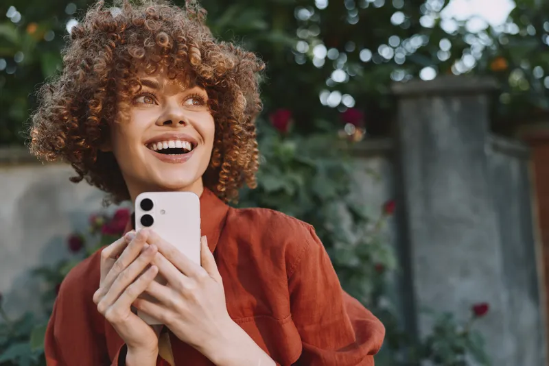 Smiling woman with curly hair holding a smartphone outdoors, bright garden background. Casual lifestyle scene, joyful expression, candid moment, natural light