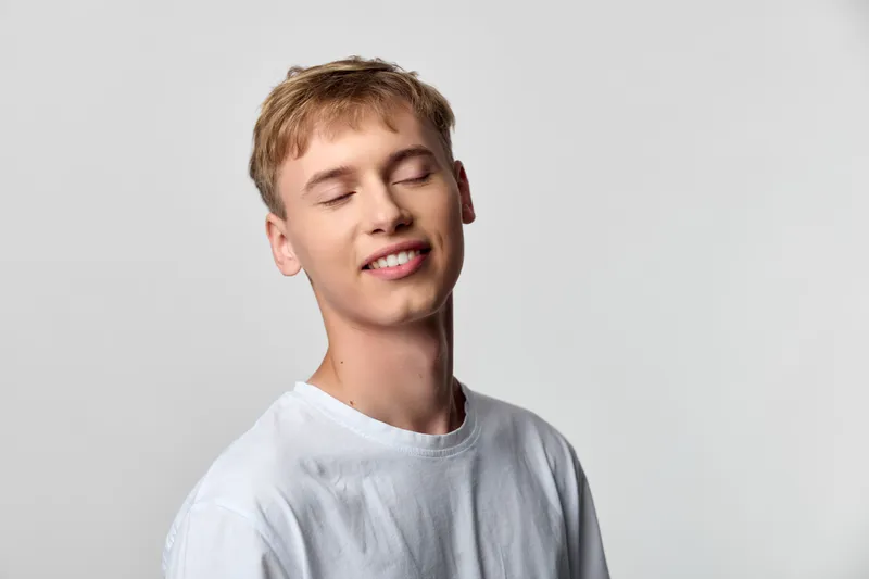 Young, relaxed man with a gentle smile wearing a white shirt in a minimal studio setting, exuding calm confidence and serene mood through soft lighting and clean background