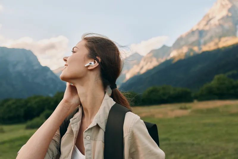 Young woman with earbuds outdoors in mountains, enjoying fresh air, casual outfit, backpack on shoulders, peaceful nature, clear sky, relaxed and happy mood
