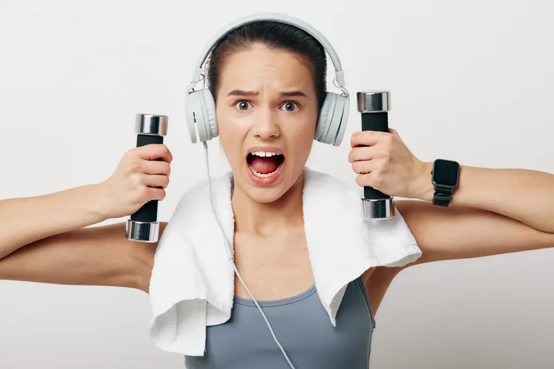 Female fitness portrait of a determined woman holding dumbbells with headphones and a towel around her neck, expressive face and sporty outfit against a clean white studio background.