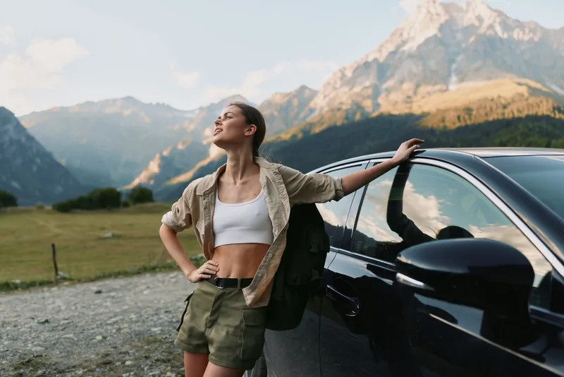 Young woman in casual outfit leans on car outdoors enjoys fresh air with mountains in background expresses freedom and lifestyle in nature travel concept.