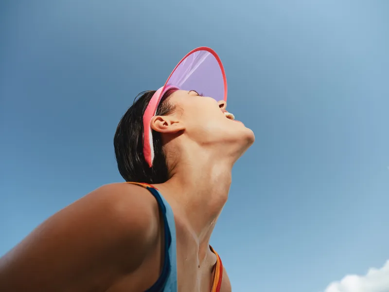 A sunlit portrait of a young athlete tilting head back beneath a translucent visor, capturing motion, energy, and brightness against a clear blue sky.