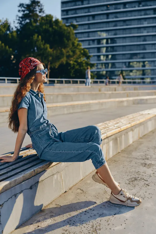 Lifestyle woman in modern boho-western denim street style wearing red bandana and sunglasses relaxing outdoors in bright film color with urban background and sunlight.
