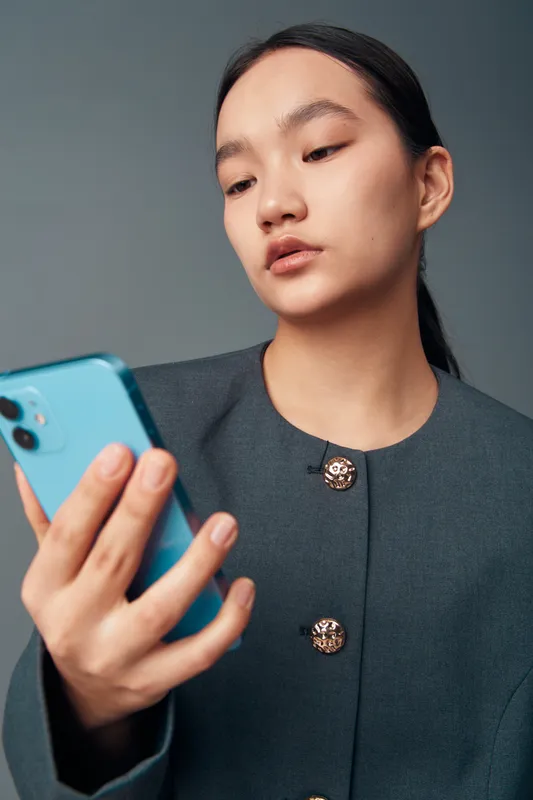 Young woman with long dark hair in grey coat looking at blue smartphone with attention and focus. Female person using mobile phone for communication or social media on neutral background.