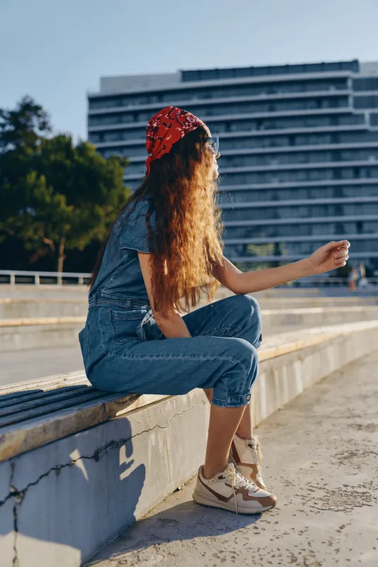 young woman in denim outfit and red bandana sitting outdoors in urban area during daytime, casual style with relaxed posture and natural light