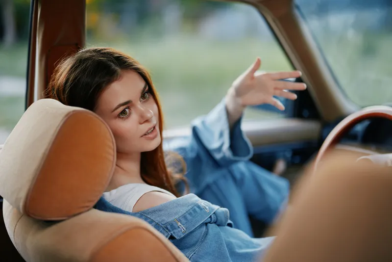 Young woman in denim jacket sitting in a vintage car, expressing joy and confidence in a sunny outdoor setting with a soft focus background.