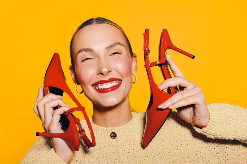 Happy woman with red lipstick holds red high heel shoes near face, smiling with closed eyes on bright yellow background. Concept of fashion, style, beauty, and joyful emotions.