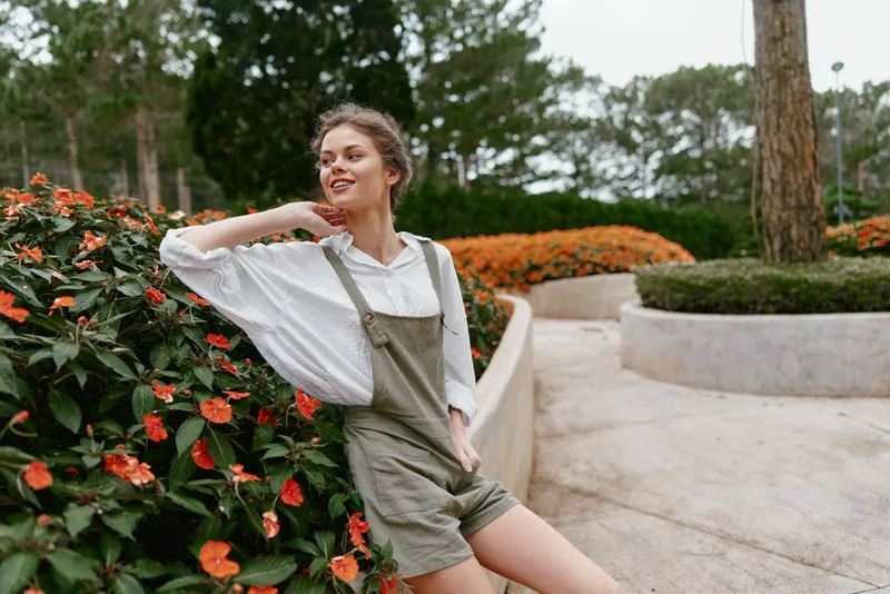 Young woman in stylish overalls smiles while posing near vibrant orange flowers in a relaxed outdoor setting, showcasing spring fashion and natural beauty