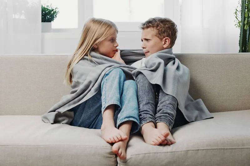 Two children wrapped in a gray blanket sit barefoot on a beige sofa facing each other with serious expressions. Natural light from the window brightens the cozy living room scene.
