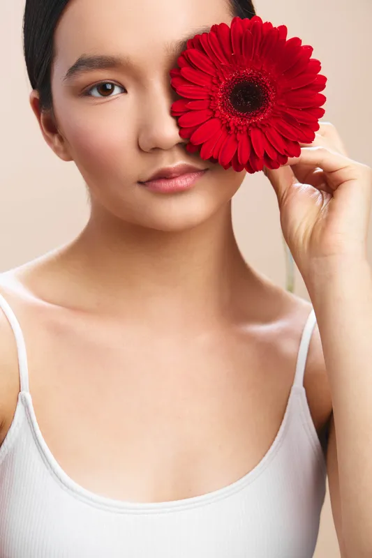 Asian woman holds a bold red flower beside her eye, creating a striking closeup with smooth skin and a calm, intimate expression in a clean studio setting.