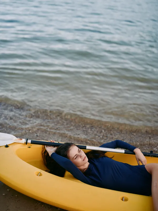 A person reclines inside a bright yellow kayak along a sandy shore, with calm water and gentle ripples behind, presenting a peaceful, minimalist composition.