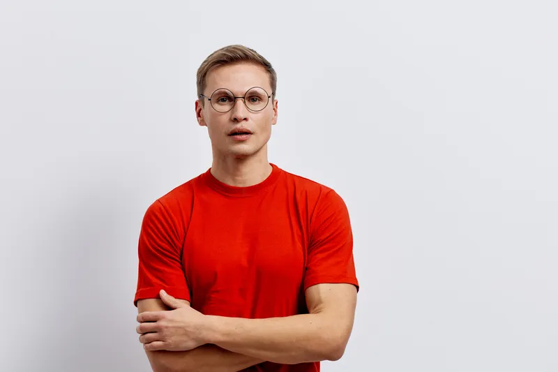 Confident young man in red t-shirt and glasses standing with arms crossed on plain white background, looking directly at camera. Studio portrait with neutral expression. People lifestyle concept