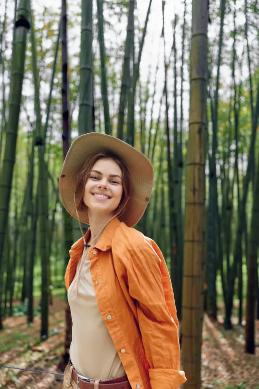 Woman smile in bamboo forest wearing hat and orange jacket, portrait outdoors in nature. Happy young traveler, casual fashion and joyful expression among tall trees and greenery.