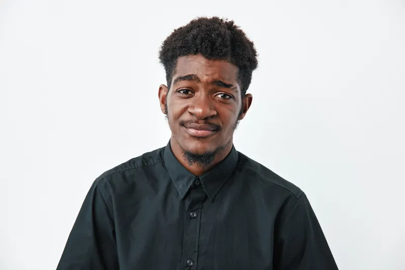 Young black man with dark skin and curly hair wearing black shirt looking at camera with uncertain expression on white background. Portrait of African ethnicity male with natural hairstyle.