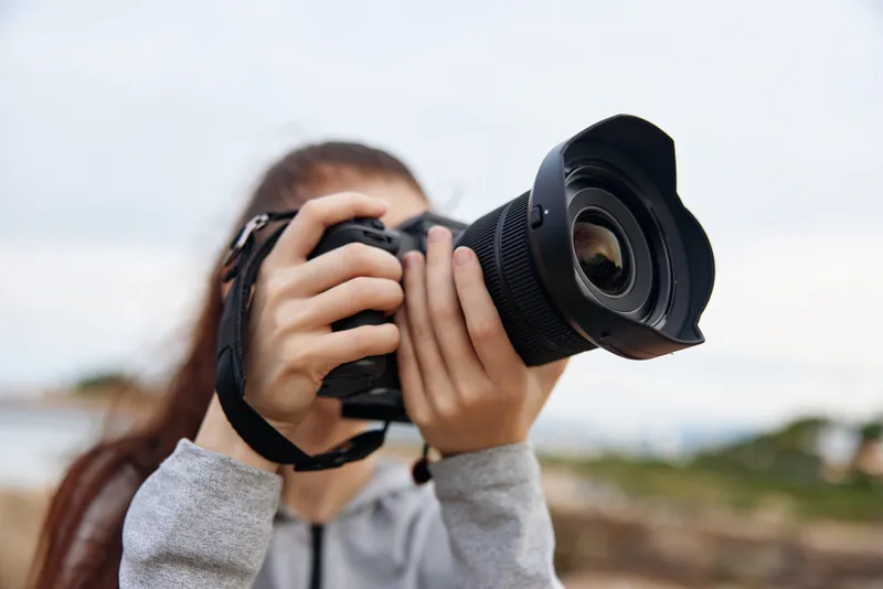 Young woman photography enthusiast capturing moments outdoors with a DSLR camera, wearing a casual gray hoodie, expressing joy amidst a scenic backdrop
