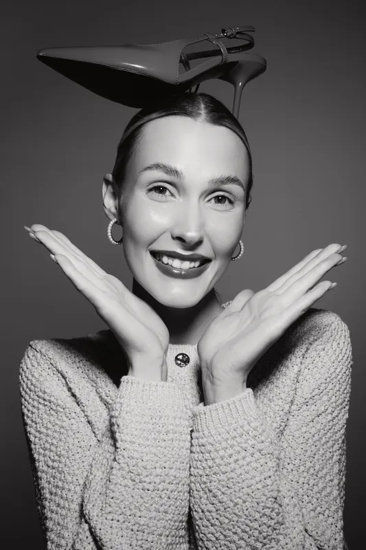 Happy Caucasian woman with fair skin and earrings smiles with crossed hands near face, wearing knitted sweater and balancing high heel shoe on head.