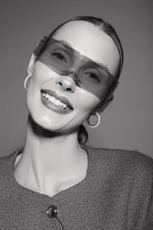 Smiling white woman with pale skin wearing transparent sunglasses and hoop earrings in studio with textured clothing.