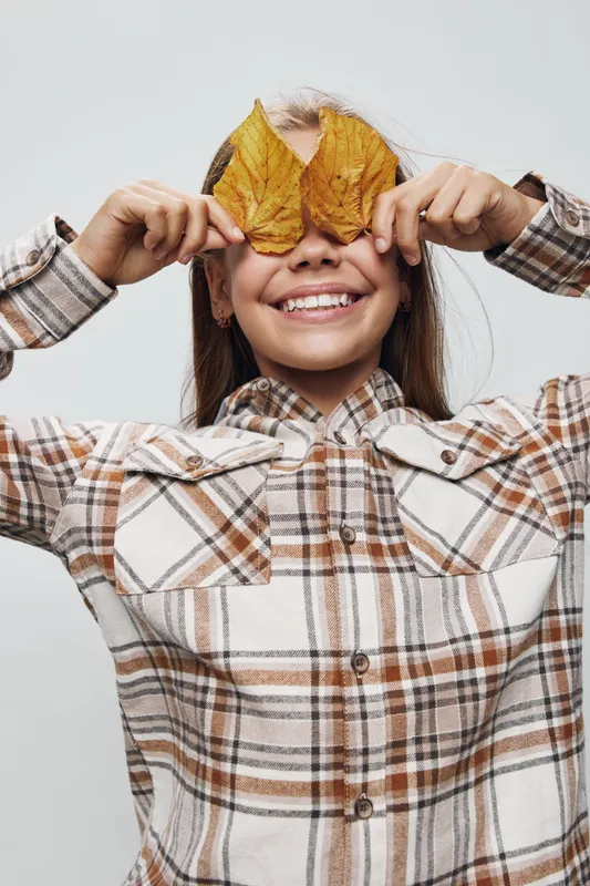 Girl smiling and holding two dry autumn leaves in front of her eyes for playful fun, childhood and seasonal mood with plaid shirt on white background indoors.