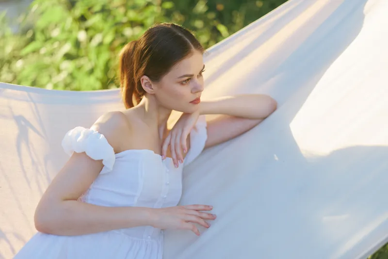 Young woman in white dress gazing thoughtfully while leaning against a soft fabric backdrop in a serene outdoor setting, evoking a sense of tranquility and reflection