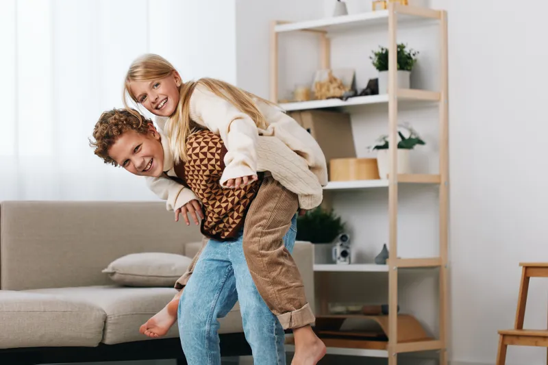 Two adults share a playful piggyback ride in a bright living room, capturing spontaneous joy and warm interaction amid cozy decor and soft natural light.
