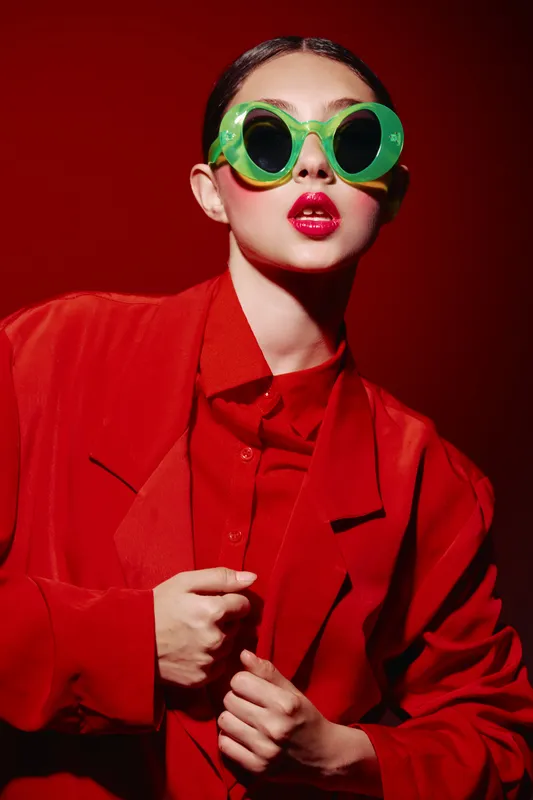 Fashionable young woman wearing bright green oversized sunglasses and red outfit looks confidently at the camera with glossy lipstick on red background. Concept of beauty and fashion accessories.