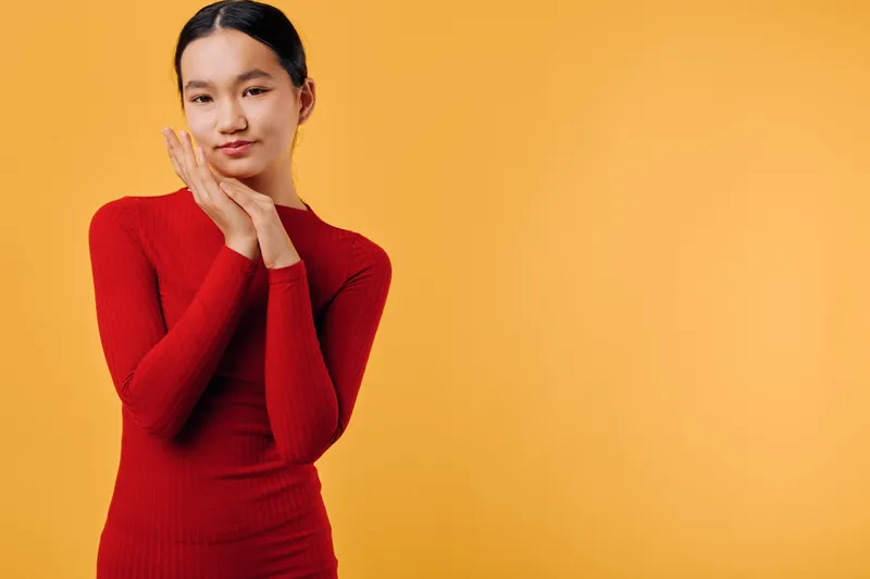Young woman in red sweater with hands near face posing against orange background. Portrait of confident female model with natural makeup and long black hair styled back.
