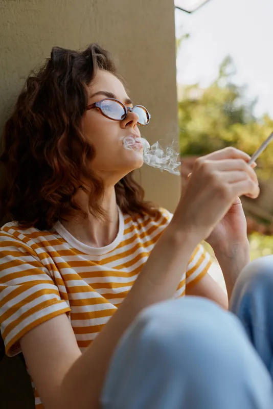Relaxed woman with curly hair wearing a striped shirt sits outdoors, smoking a cigarette in a casual moment. Warm natural light, soft shadows, and a calm mood of everyday lifestyle.