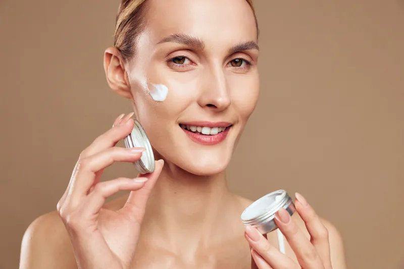 Smiling woman applying white face cream on cheek holding jar with beige background. Concept of skincare routine, beauty care, wellness, and natural skin treatment in soft lighting.