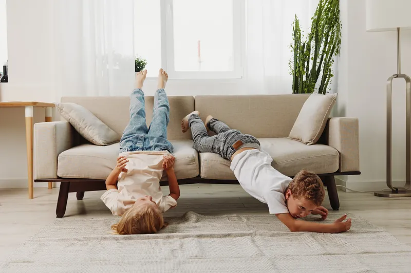 Two playful children lie upside down on a beige carpet in a bright living room with a modern sofa and green plants. Concept of childhood fun and home comfort.