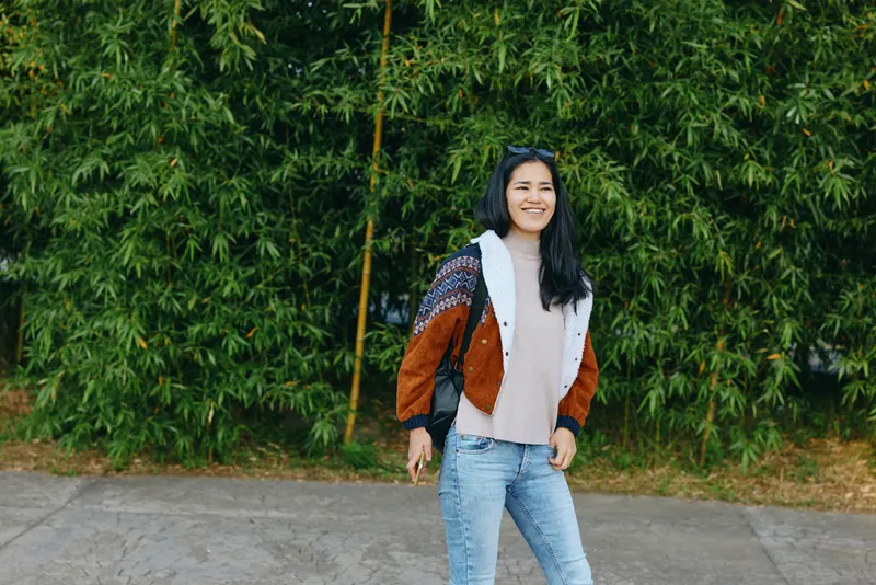 Smiling woman stands on a sidewalk beside a lush green hedge, wearing a color block jacket and denim jeans, capturing a casual street fashion moment with relaxed style and sunny outdoor vibe.