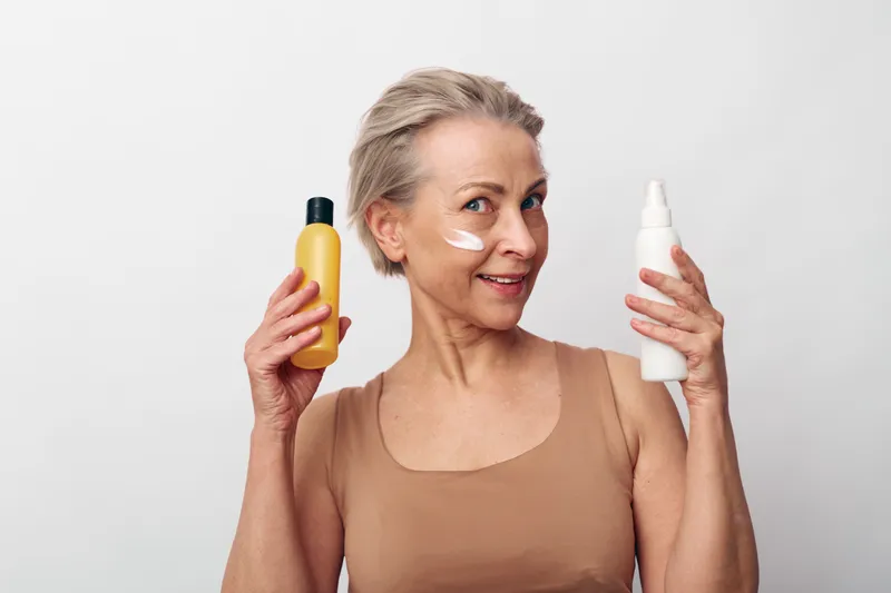 Mature woman with short hair applies cream on cheek while holding two cosmetic bottles, smiling in beige top on white background. Concept of skincare, beauty, self-care, and anti-aging routine.