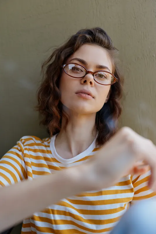 Smiling woman with short brown hair wearing a striped yellow shirt and glasses indoors. Casual lifestyle at home, self care vibe. Natural light creates a warm, relaxed, confident mood.
