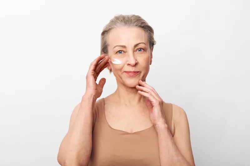 Mature woman with short grey hair applies face cream on cheek, smiling gently against plain white background. Concept of skincare, aging gracefully, self-care, and natural beauty.