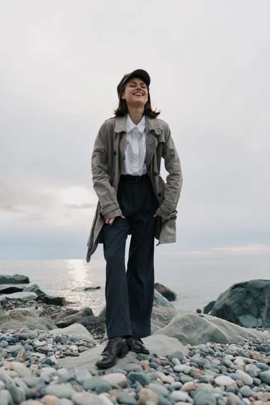 Young woman in casual outfit and cap stands on rocky beach smiling happily. Overcast sky and calm sea create relaxed natural atmosphere. Concept of outdoor lifestyle and joy.