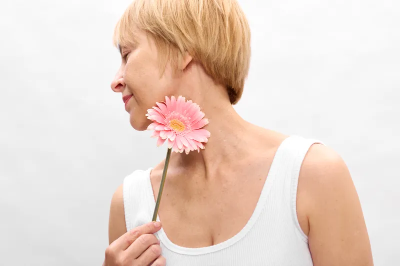 Woman in closeup holding a pink gerbera flower near her face, blonde hair in side portrait, soft smile, calm mood, white background, minimalist studio light, fresh look