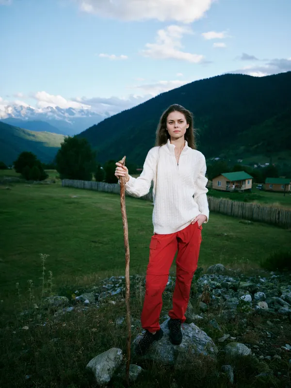 Outdoor hiking scene with a woman standing on rocky ground holding a wooden staff, wearing red pants and a white sweater, in a green valley with distant mountains and blue sky.