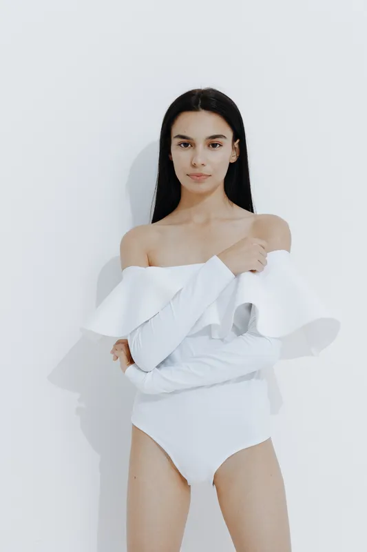 slim woman sport fitness wearing white off-shoulder bodysuit posing confidently with straight black hair and minimal makeup against a bright plain background.