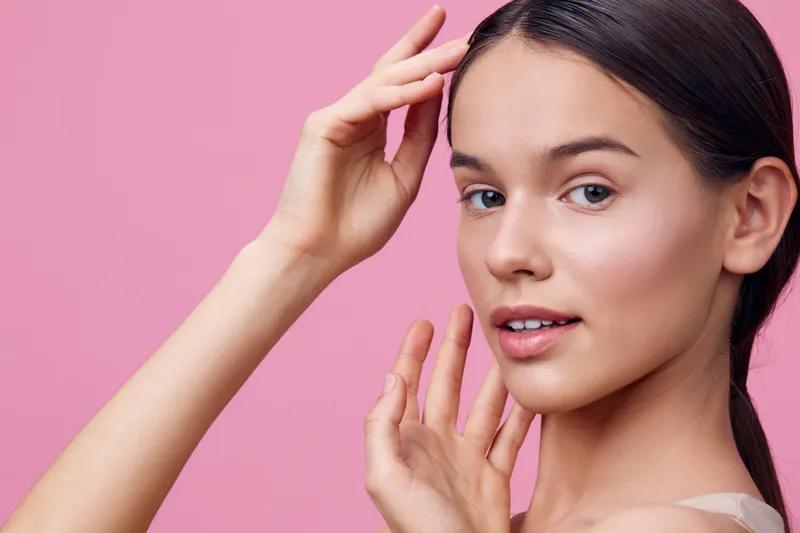 Young woman with perfect skin showcasing skincare routine against a soft pink background, emphasizing beauty and confidence