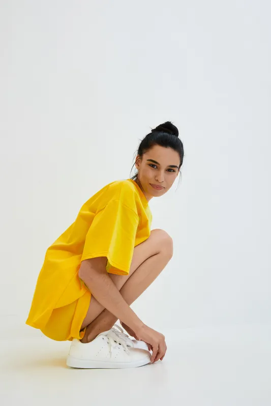 young woman in yellow oversized dress crouching, wearing white sneakers, minimalistic background, casual style, natural light, indoor setting
