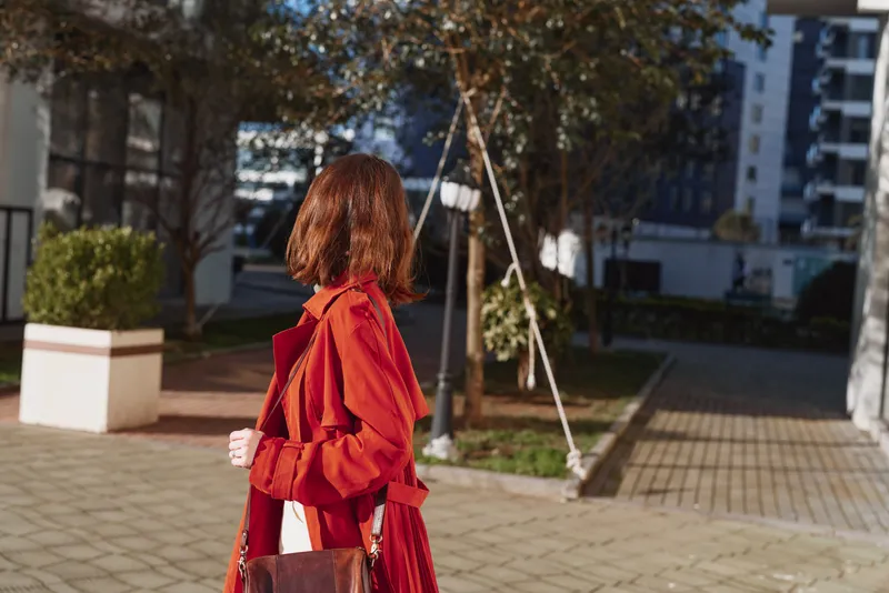 A woman in a bright red coat walks through a sunny urban plaza, adjusting her jacket with a confident stride while a bag rests at her side, conveying purposeful style and solo presence.