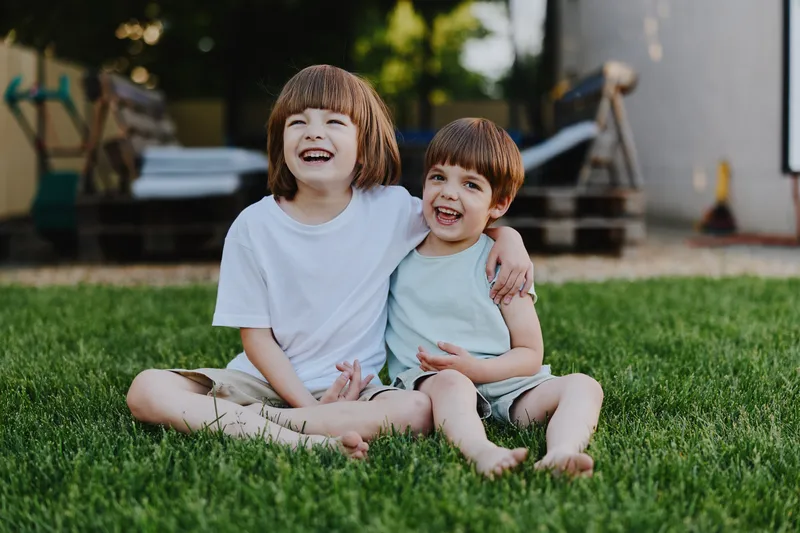 Two happy boys sitting on green grass, sharing laughter and joy. They wear casual summer clothes and have bright smiles, showcasing a playful and carefree moment.