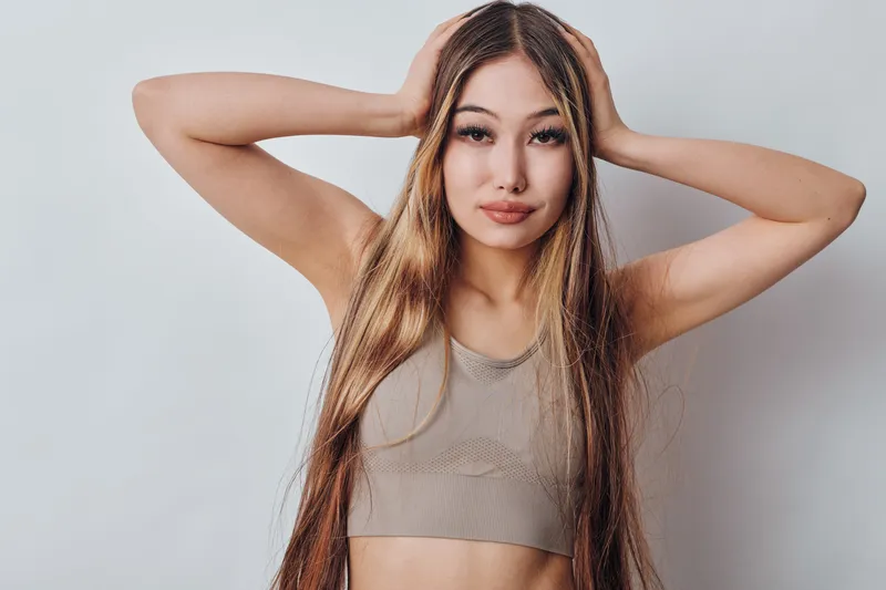 Young woman with long hair wearing sleeveless top posing with hands on head in studio. Portrait, beauty, fashion and confident expression with casual style on neutral background.