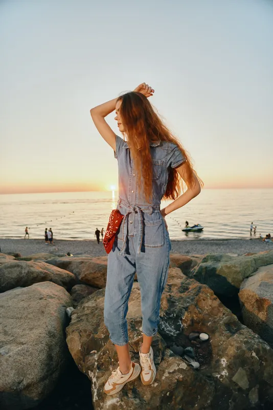 lifestyle woman in modern boho-western denim street style stands on rocks at sunset, film color capturing relaxed seaside fashion mood