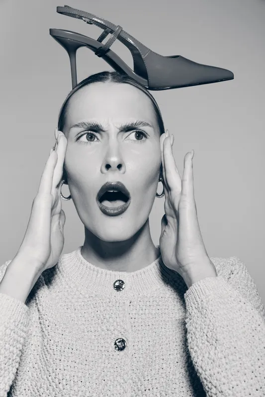 White woman with fair skin expresses surprise holding head with hands. A high heel shoe balanced on her head creates unusual fashion concept and expressive portrait.