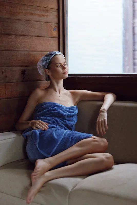 young woman wrapped in blue towel relaxing indoors beside window, natural light illuminating calm expression, wooden wall background creating cozy atmosphere