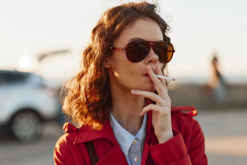 Young woman with curly hair wearing sunglasses and red coat smoking cigarette outdoors during golden hour urban background casual style