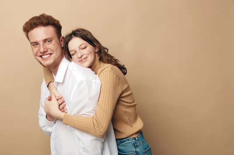 Smiling young couple embraces happily against beige background. Woman in brown sweater hugs man in white shirt. Concept of love, affection, relationship, and casual lifestyle.